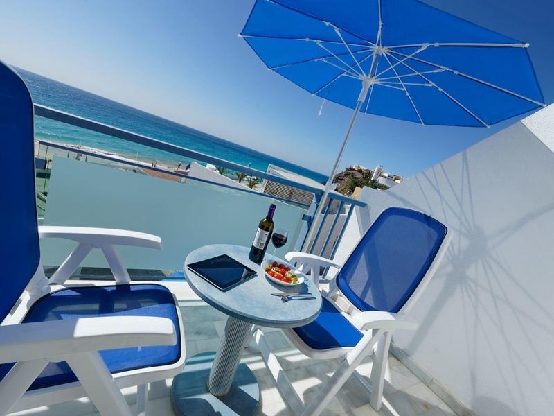 Balcony with blue chairs, table with wine, and umbrella overlooking a waterfront view.
