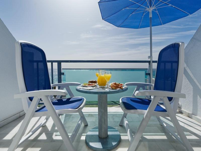 Two chairs and a table with drinks on a balcony overlooking the sea with a blue umbrella.