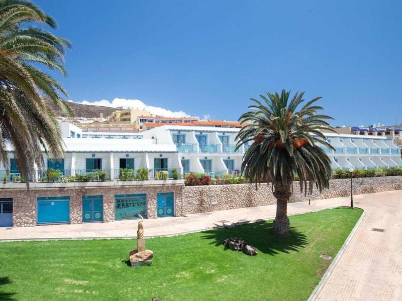 Coastal hotel with white walls, blue shutters, palm trees, and clear blue sky.