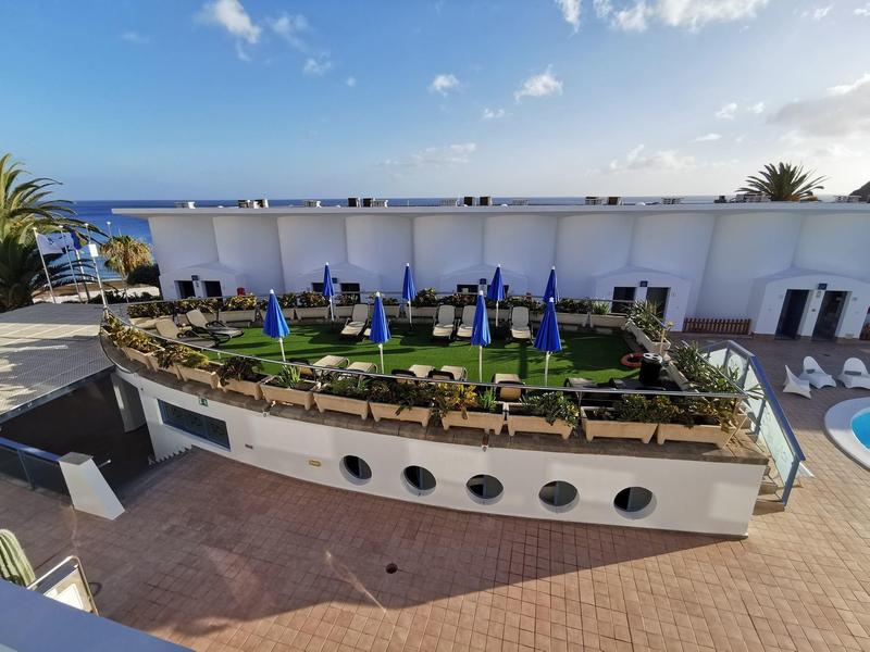Outdoor rooftop lounge area with green grass, blue umbrellas, and white seating under a clear sky.