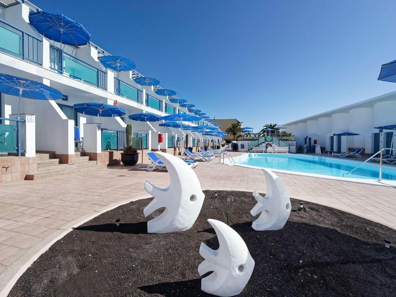 Outdoor pool area at a hotel with sun loungers, blue umbrellas, and white shark fin sculptures.