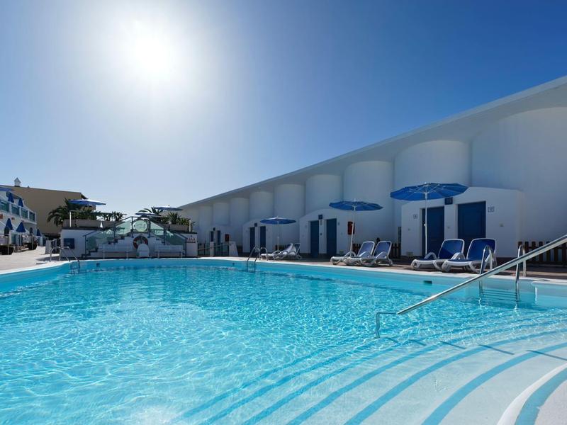 Sunny outdoor pool with lounge chairs and blue umbrellas at a modern hotel.