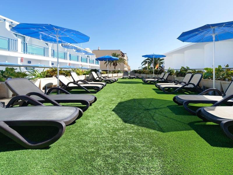 Outdoor lounge area with black sunbeds and blue umbrellas on green grass under blue sky.