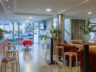 Lounge di hotel luminosa con sgabelli da bar, piante e area salotto con grandi finestre.
