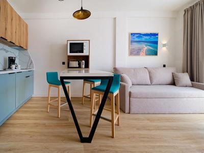 Moderner Wohnbereich mit Küchenzeile, Esstisch, zwei Stühlen und Sofa neben großem Fenster.