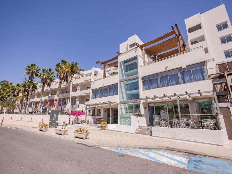 Moderno edificio dell'hotel con terrazze soleggiate e palme lungo una strada costiera soleggiata.