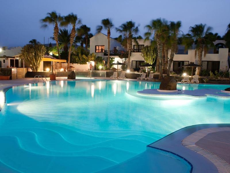 Piscina dell'hotel illuminata con palme e camere al crepuscolo.