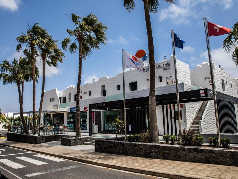 Edificio moderno bianco di un hotel con bandiere e palme lungo una strada soleggiata.