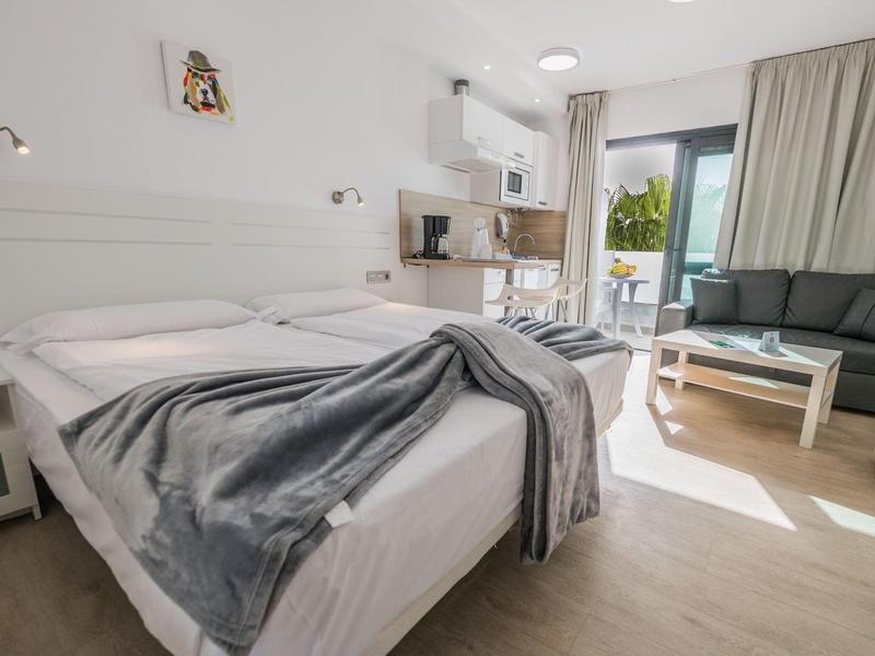 Camera d'hotel moderna con letto matrimoniale, divano, angolo cottura e balcone con luce naturale.