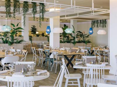Modern restaurant with white wooden chairs, round tables, hanging plants, and blue pendant lights.