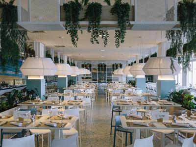 Bright, spacious hotel restaurant with white tables, chairs, hanging plants, and large windows.