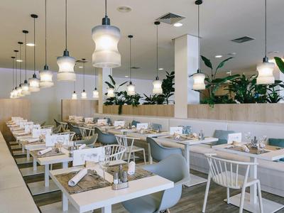 Modern hotel restaurant with white tables, chairs, blue cushions, and hanging lights.
