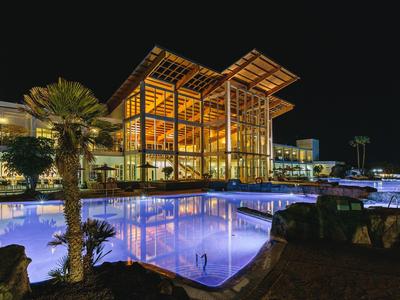 Beleuchtetes Hotelgebäude mit großen Glasfronten neben einem Pool bei Nacht.
