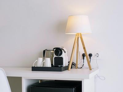 Minimal hotel desk setup with coffee maker, mugs, and a tripod lamp in soft lighting.