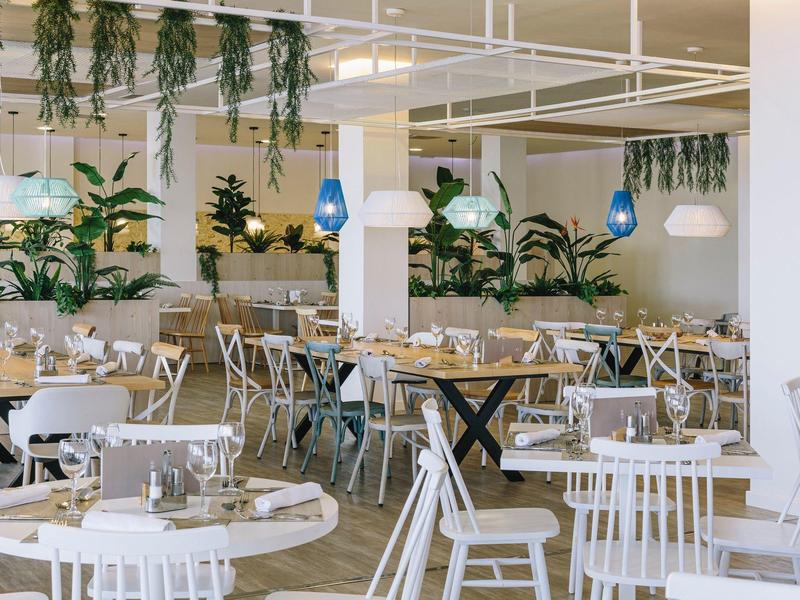 Modern restaurant with white wooden chairs, round tables, hanging plants, and blue pendant lights.