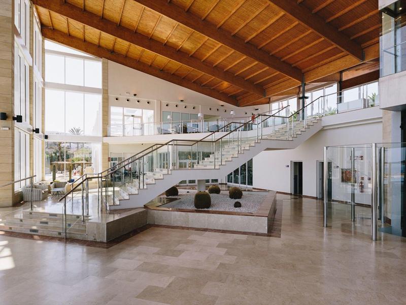Spacious hotel lobby with glass staircase, wooden ceiling, and large windows.