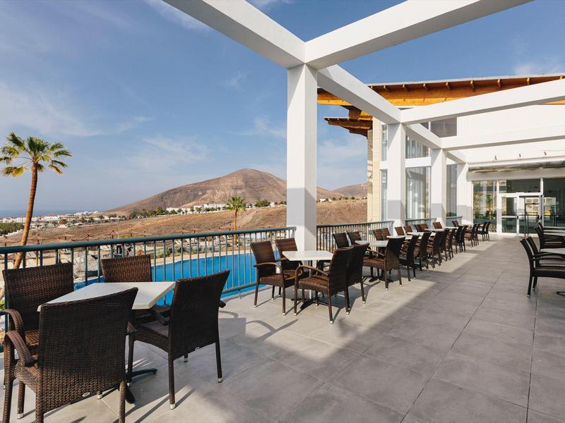 Outdoor terrace with tables and chairs overlooking a pool and mountain scenery.