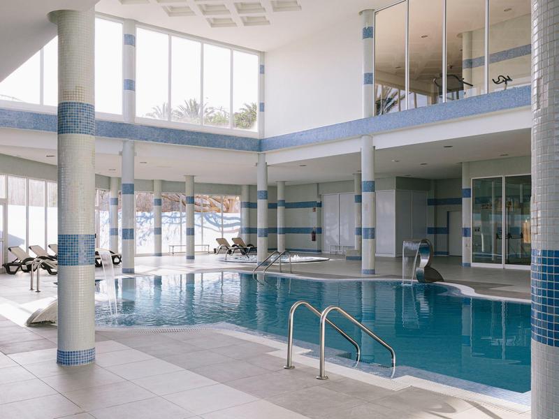 Indoor hotel swimming pool with blue tiles and glass windows letting in natural light.