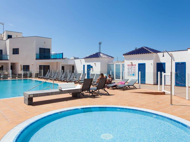 Hotel pool area with multiple pools, sun loungers, and modern white buildings under clear sky.