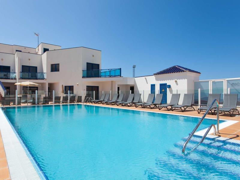 Modern outdoor pool with sun loungers in front of white hotel building under clear blue sky.