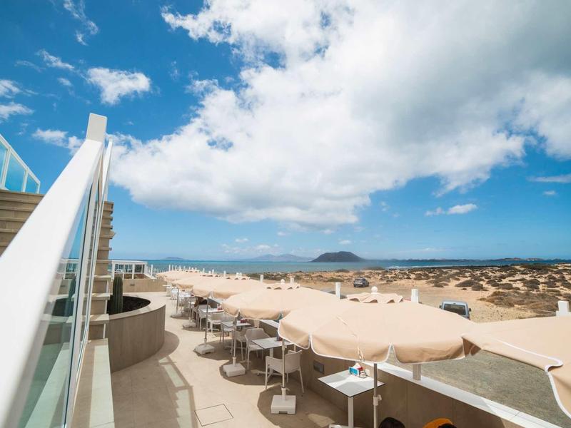 Area terrazza con tavoli e ombrelloni con vista sul paesaggio e sul mare sotto un cielo azzurro.