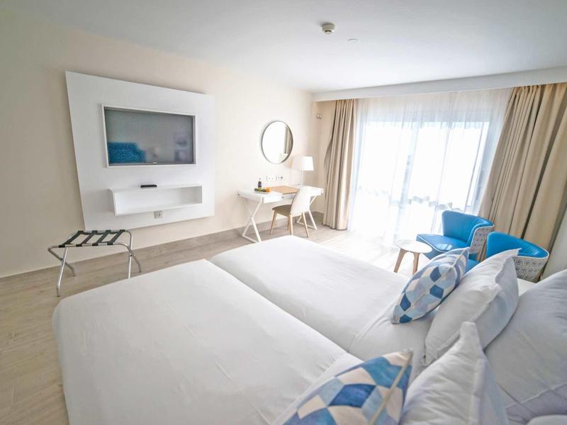 Camera d'hotel luminosa con due letti, scrivania, sedia e balcone con luce naturale.