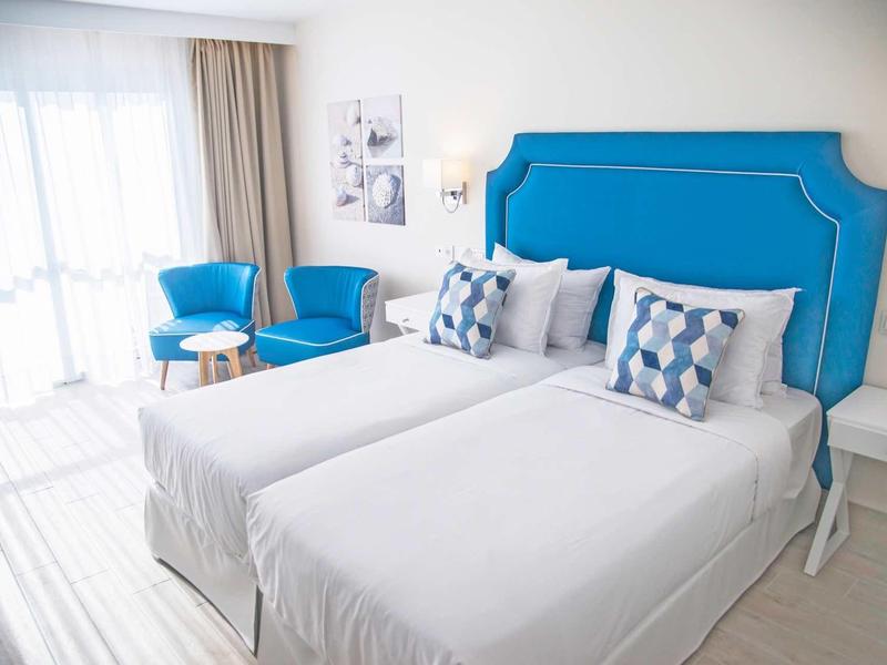 Camera d'hotel luminosa con due letti singoli, testiera blu e due poltrone blu vicino alla finestra.