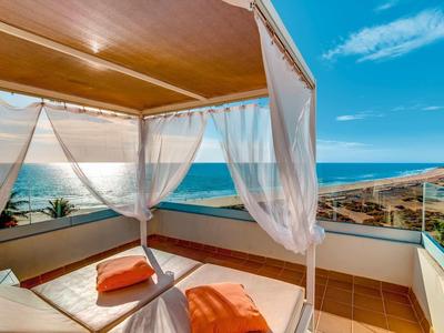 Terrasse mit Bett und weißen Vorhängen, Blick auf türkisfarbenes Meer und klaren Himmel.