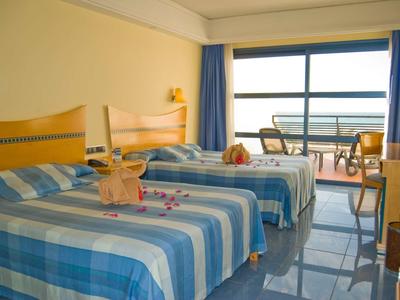 Camera d'hotel con due letti, biancheria a righe blu e balcone con vista mare.