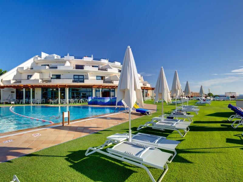 Hotel con grande piscina all'aperto, lettini e ombrelloni su prato verde sotto un cielo azzurro.
