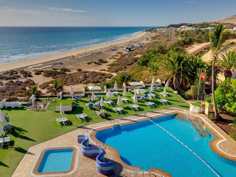 Area piscina con ombrelloni e lettini, spiaggia e mare sullo sfondo sotto un cielo azzurro.
