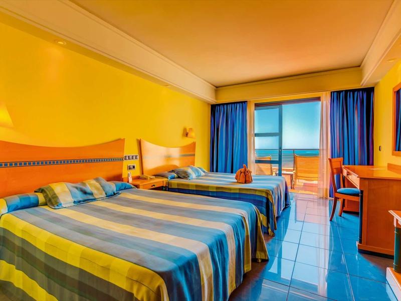 Camera d'hotel con due letti, pareti gialle e vista sul mare attraverso grandi finestre.