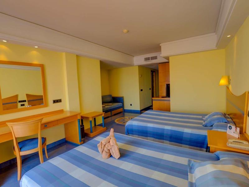 Camera d'hotel con due letti singoli, scrivania, specchio e zona salotto in tonalità giallo-blu.