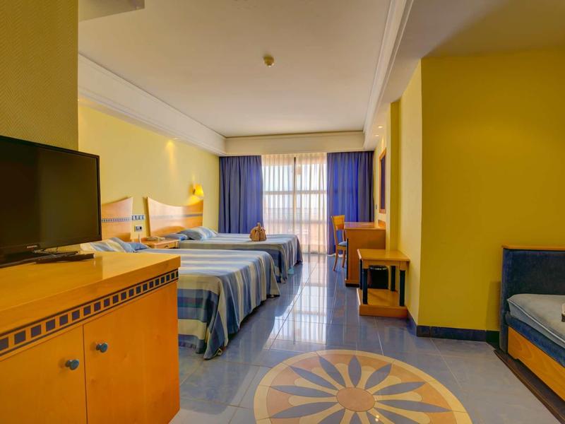 Camera d'hotel luminosa con due letti, TV, zona salotto e vista sul balcone.