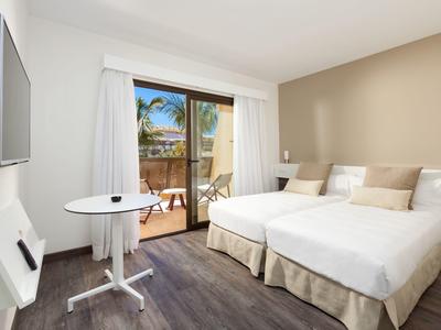 Camera d'hotel moderna con letto matrimoniale, tavolo rotondo e balcone con vista su palme.