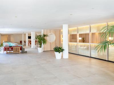 Ingresso di hotel luminoso con pavimento in piastrelle grigie, piante verdi e banco reception in legno.