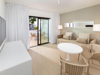 Bright hotel room with seating area, sofa, coffee table, and balcony with outdoor view.