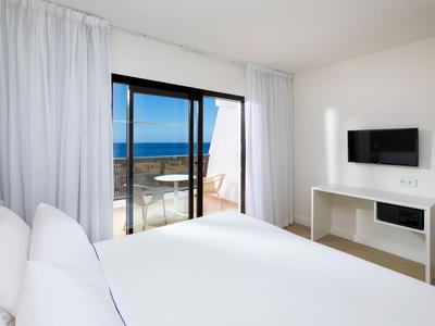 Camera d'hotel moderna con letto bianco, TV a parete e vista mare dalla porta finestra.