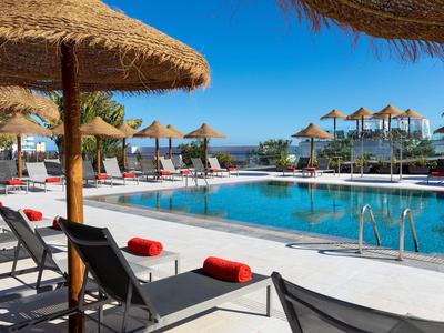 Piscina di hotel soleggiata con lettini e ombrelloni di paglia.