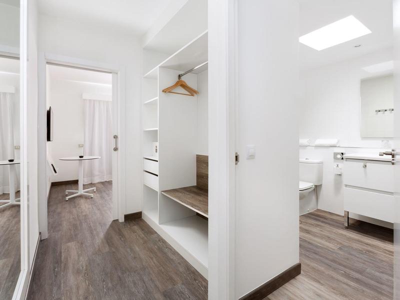 Camera d'hotel moderna con pavimento in legno, armadio aperto, specchio e bagno luminoso.