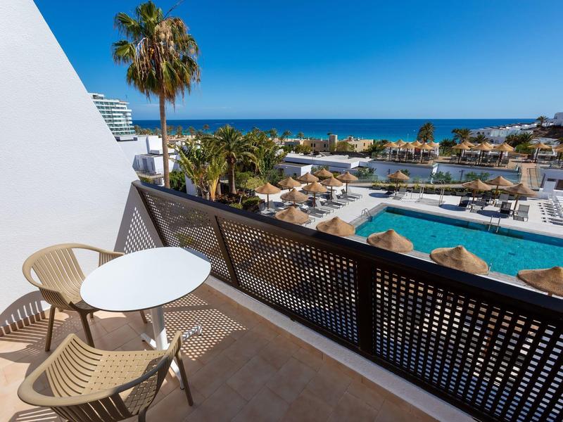 Balcone dell'hotel con tavolo, sedie, piscina, palme e vista sul mare.