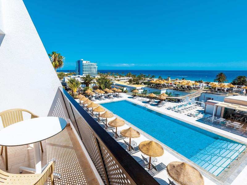 Vista su piscina all'aperto di un hotel con terrazza e sedie di fronte al mare sotto un cielo blu.