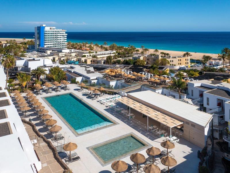 Vista di una località balneare con piscine, ombrelloni, edifici bianchi e spiaggia sullo sfondo.