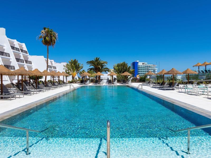 Piscina esterna di un hotel con lettini e ombrelloni sotto un cielo azzurro.