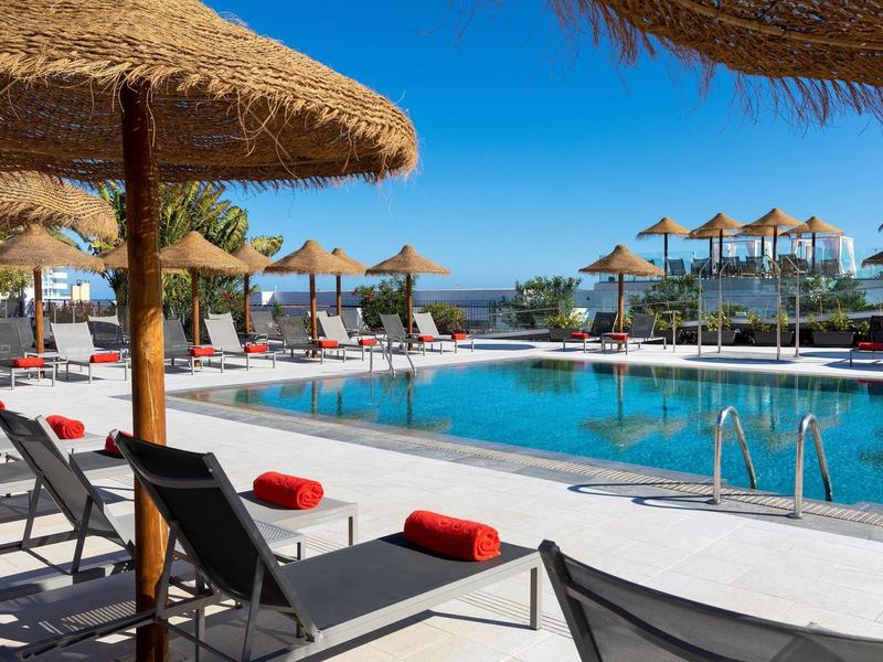 Piscina di hotel soleggiata con lettini e ombrelloni di paglia.