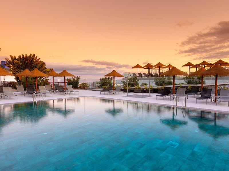 Piscina esterna luminosa con ombrelloni di paglia al tramonto.