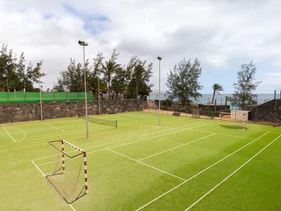 Leerer Tennisplatz mit grünem Kunstrasen, umgeben von Bäumen und beleuchtet durch hohe Lampen.