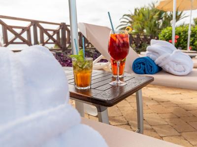 Zwei Relaxliegen mit Handtüchern und zwei bunte Cocktails auf einem Holzbeistelltisch im Freien.