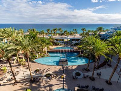 Blick auf einen blauen Pool mit Palmen, Strand und Meer im Hintergrund bei sonnigem Wetter.