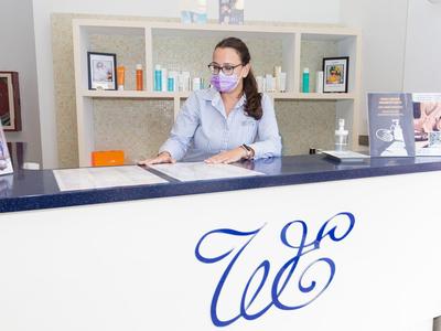 Eine Frau mit Maske sitzt hinter einem weißen Empfangstresen mit blauer Schrift in einer modernen Klinik.